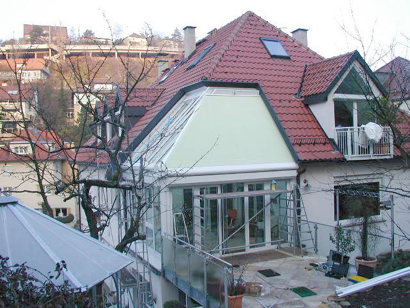 Mehrstöckiges Wohnhaus mit rotem Satteldach, großem Wintergarten und umlaufendem Balkon, umgeben von kahlen Bäumen.