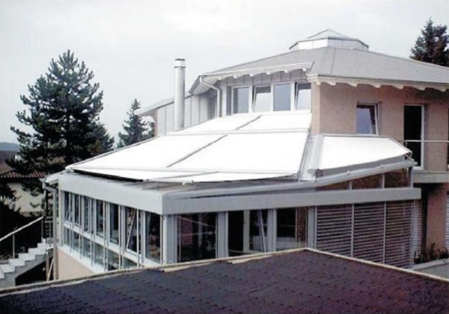 Modernes Gebäude mit großem, flachem Glasdach und umlaufenden Fenstern, umgeben von Bäumen.